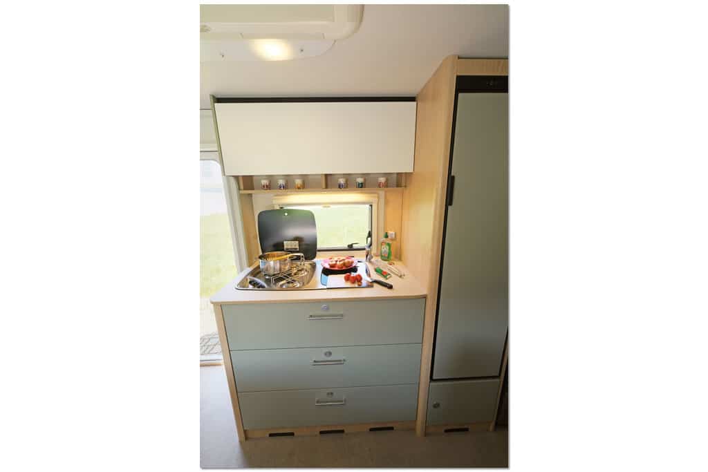 Die Küche im LMC Style neben der Eingangstür besteht aus einem Modul mit drei großen Schubladen mit salbeifarbenen Fronten, darüber Kochfeld und abdeckbares Spülbecken, ein Oberschrank mit weißer Front und neben dem Küchenmodul ein hoher Kühlschrank mit salbeigrüner Front.