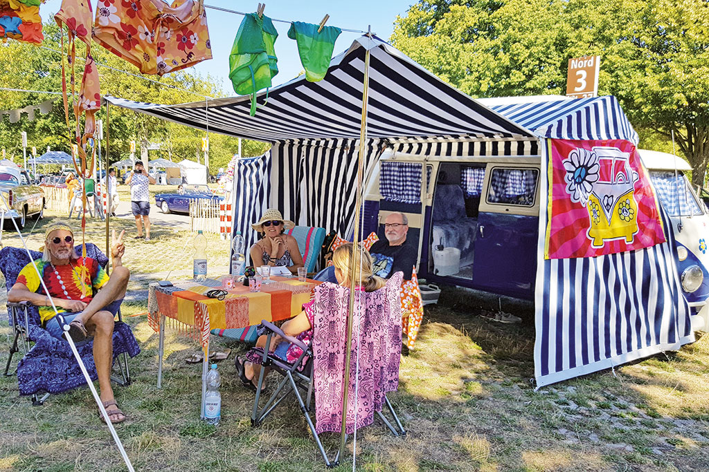 Vier als Hippies gekleidete Teilnehmer der Classic Days 2022 sitzen am Picknicktisch vor blau-weißem VW T2-Bus.