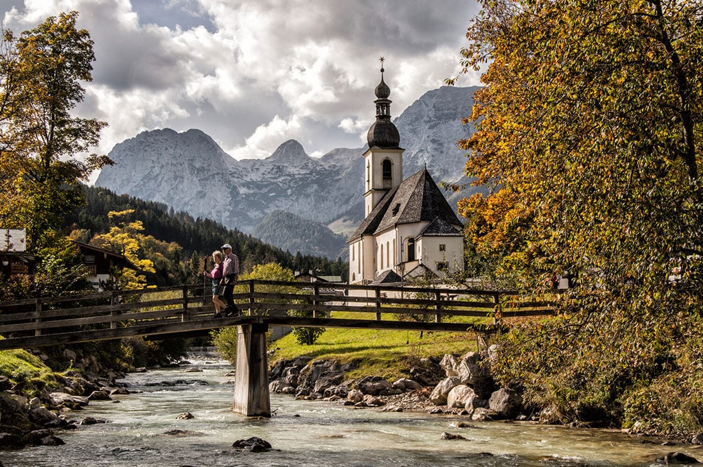 Zwei Wanderer überqueren kleinen Fluss auf schmaler Brücke, im Hintergrund eine kleine Kirche und Alpenkette