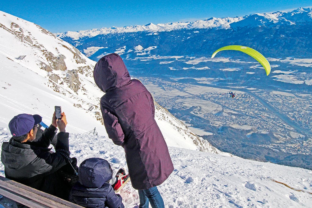 Eine Familie beobachtet einen Paraglider, der zwischen schneebedeckten Gipfeln durch die Luft gleitet