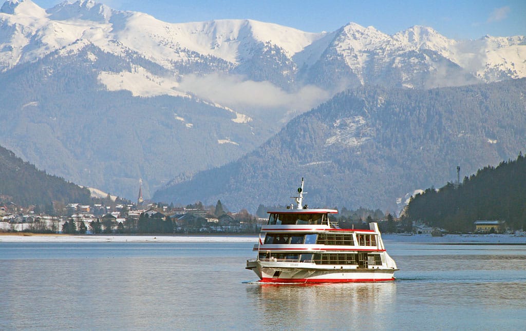 Ein Ausflugsschiff fährt auf einem See in winterlicher Berglandschaft, am Ufer im Hintergrund sieht man eine kleine Ortschaft.
