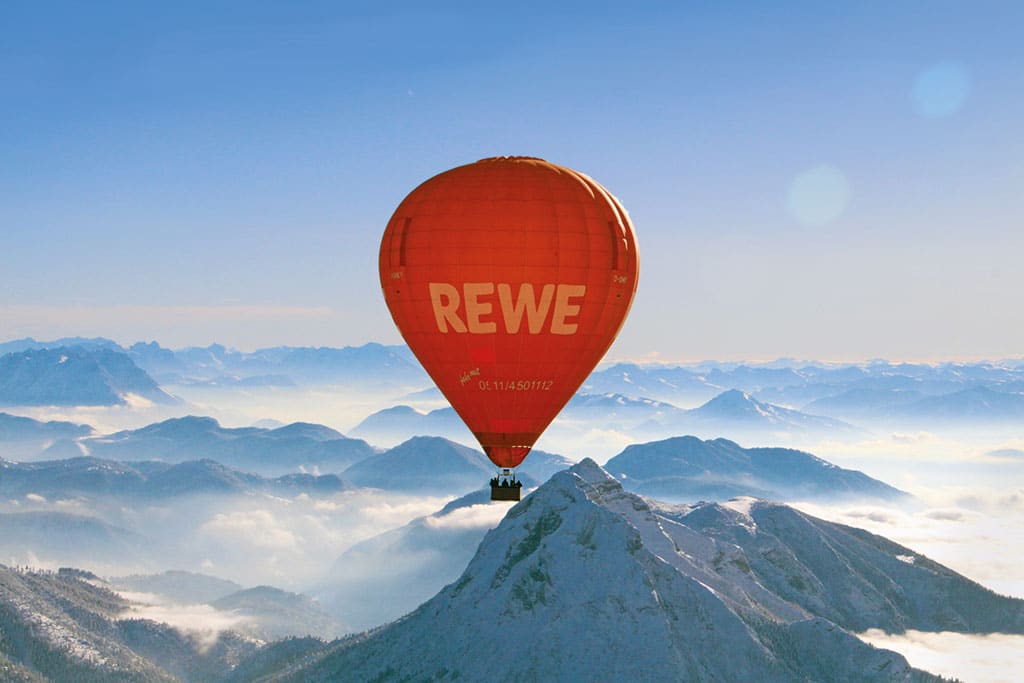 Roter Heißluftballon mit Rewe-Werbeslogan über winterlicher Berglandschaft
