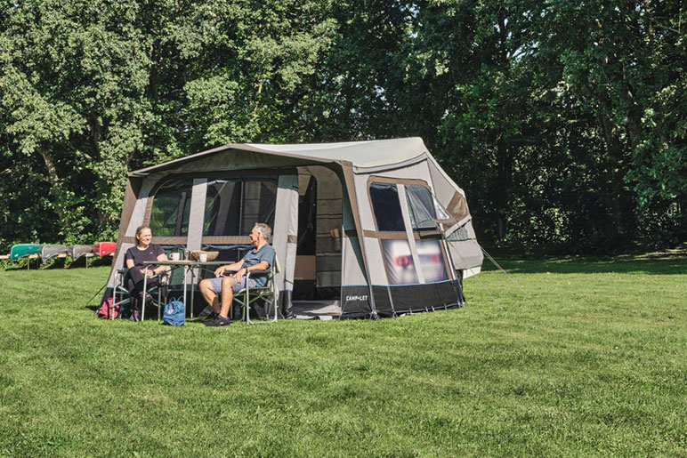 Grauer Faltcaravan und zwei davor sitzende Menschen auf einer Wiese
