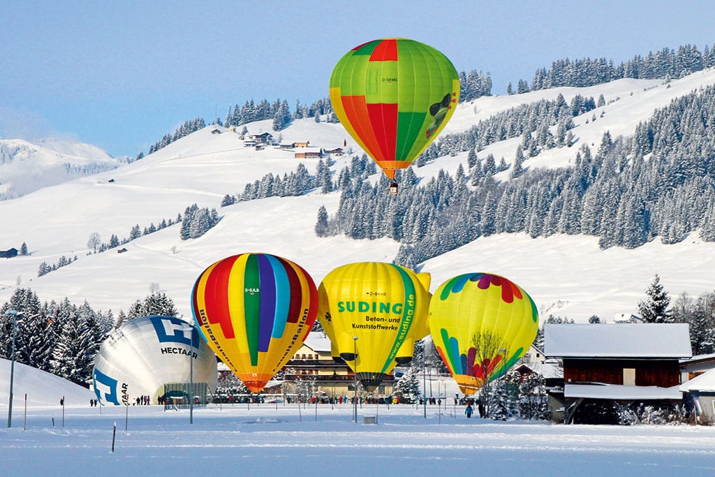 Mehrere bunte Heißluftballons beim Abheben in winterlicher Berglandschaft