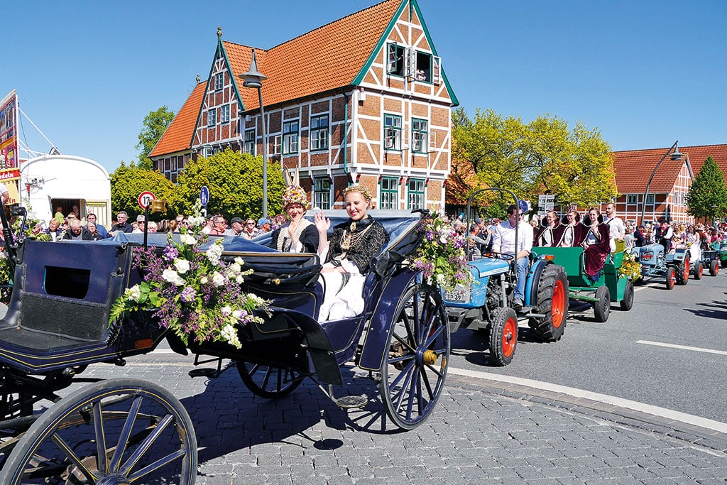Zwei junge Frauen in einer blumengeschmückten Kutsche, dahinter folgt ein Traktor sowie eine weitere Kutsche, im Hintergrund ein altes Fachwerkhaus