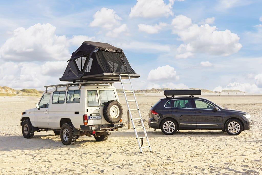 Weißer Geländewagen mit aufgestelltem Dachzelt, daneben ein dunkler PKW mit geschlossenem Dachzelt an einem weitläufigen Strand
