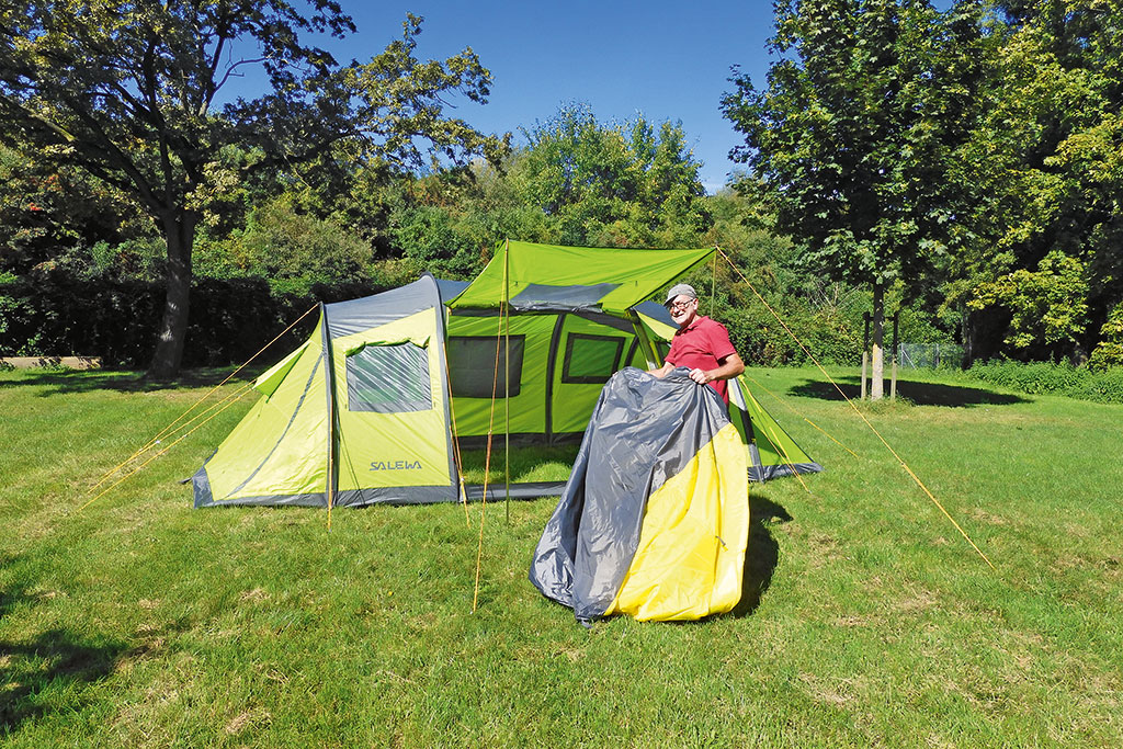 Mann trägt gelb-graue Innenzelte in auf einer Wiese aufgestelltes, hellgrün-graues Tunnelzelt