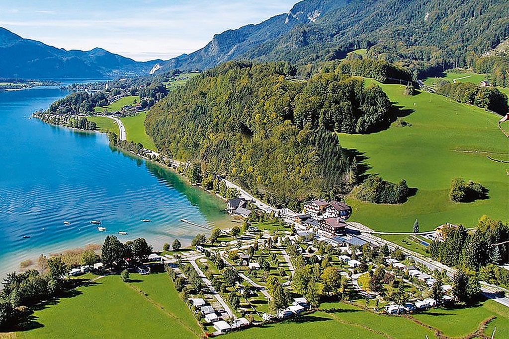 Ein kleiner Campingplatz direkt am Ufer eines Sees mit türkisblauem Wasser in einer Wiesen- und Waldlandschaft am Fuße hoher Berge