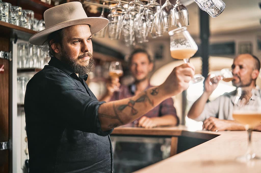 Ein Mann mit Hut hinter einem hölzernen Bartresen hält ein Glas gezaptes Bier gegens Licht, im Hintergrund stehen einige Personen am Tresen
