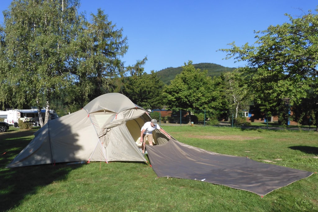 Der graue Boden für das Zelt, wird in das Zelt getragen. Das Zelt steht auf Wiese mit Bäumen im Hintergrund.