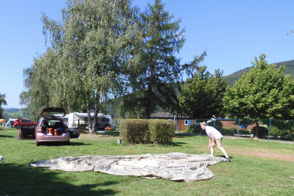 Mann legt Zelt auf Zeltboden auf, auf grüner Wiese. Bäume, Hecken und Autos im Hintergrund.