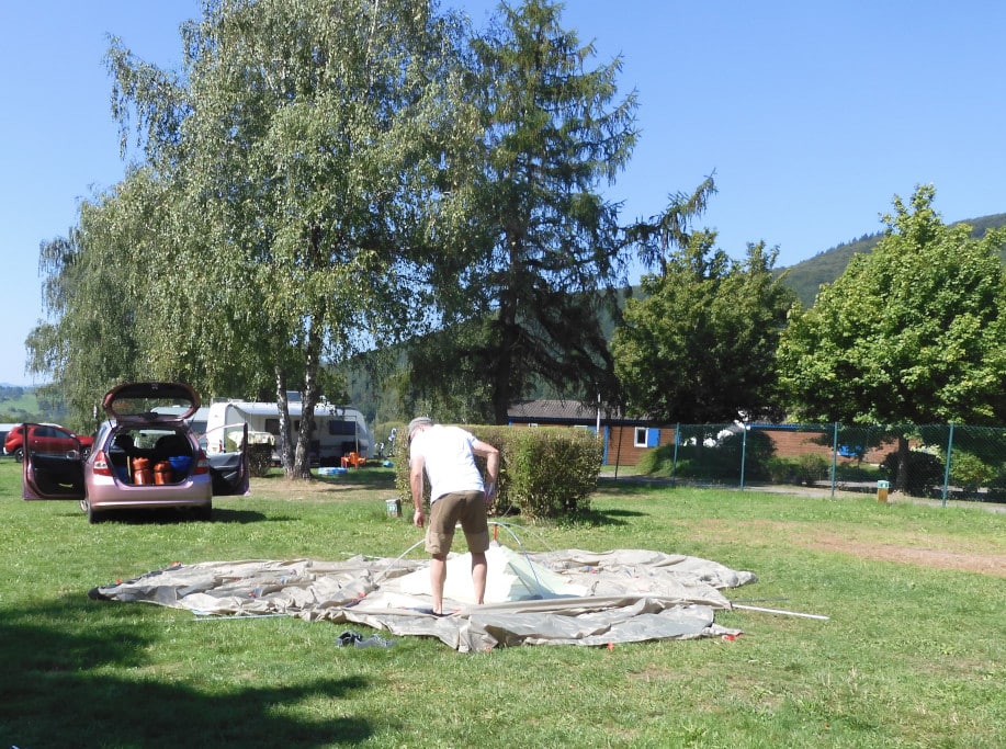 Das Kuppeldreieck des Zeltes wird durch Mann auf grüner Wiese aufgebaut. Baüme, Hecken im Hintergrund.