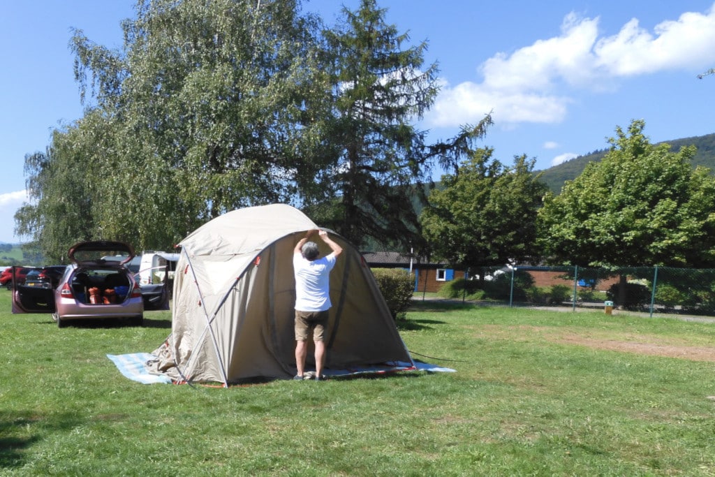 Mann montiert Abdeckungen am Zelt. Auf Wiese, mit Hecken im Hintergrund.