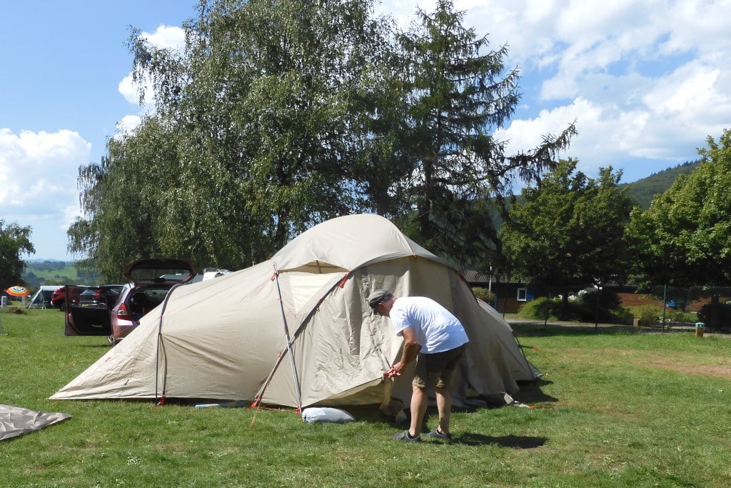 Mann fixiert Eingang des Zeltes. Auf Wiese und Bäume im Hintergrund.