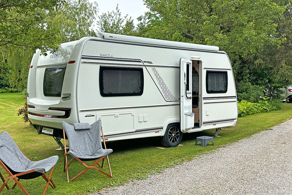 Wohnwagen auf einer Wiesenparzelle, dahinter ein paar Bäume, vor dem Wohnwagen stehen zwei Campingstühle