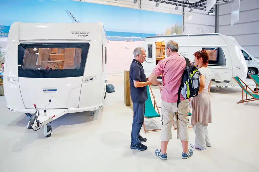 Drei Menschen unterhalten sich in einer Messehalle, im Hintergrund stehen einige Wohnwagen