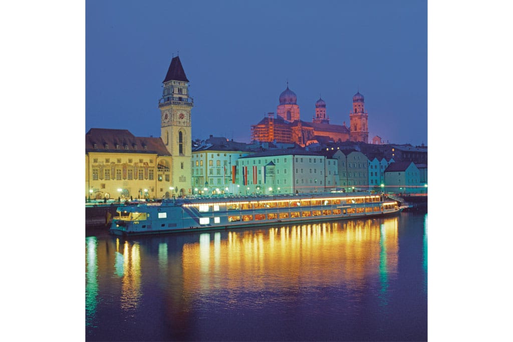 Historische Gemäuer mit einem hohen Turm am Ufer eines Flusses bei Nacht, im Hintergrund ein altes Schloss, eun Ausflugsschiff fährt auf dem Wasser