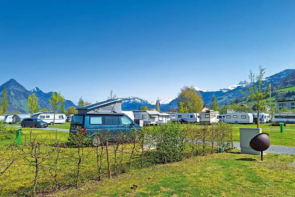 Campingfahrzeuge auf einer großen Wiese eines Campingplatzes, am Horizont eine Kette schneebedeckter Berge