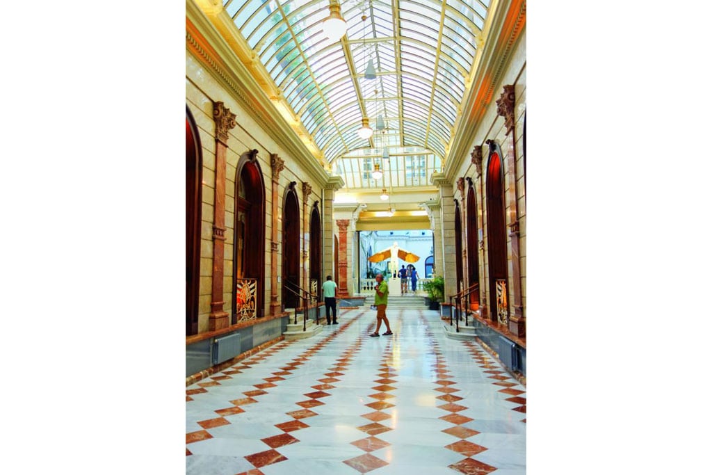 Eine langgestreckte, sehr hohe Halle mit Torbögen an jeder Seite, das gewöbte Dach besteht aus zahlreichen Fenstern, der Boden aus weißem und braunorangefarbenen quadratischen Steinen.