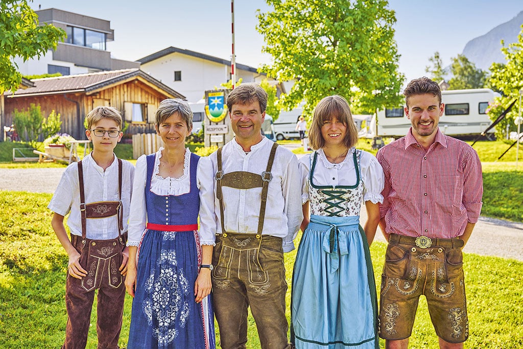 Eine Frau und ein Mädchen in blauen Dirndln, ein Mann mittleren Alters, ein junger Mann und ein Junge in Lederhosen vor einigen Gebäuden und Campingfahrzeugen auf einem Campingplatz