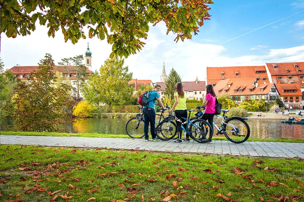 Drei Radfahrer neben ihren Fahrrädern an einem Flussufer, auf der anderen Seite des Flusses befinden sich einige Gebäude. Einige Laubbäume mit herbslich bunt gefärbten Flättern säumen die Flussufer.