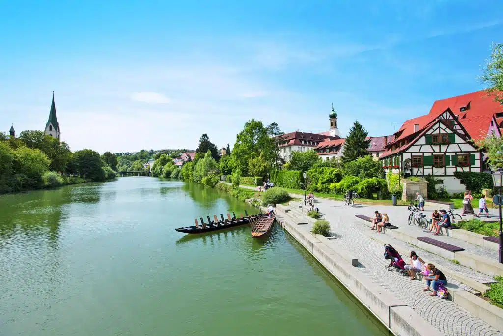 Einige Menschen sitzen auf einer verbreiterten Promenade an einem Flussufer, hinter ihnen stehen einige Gebäude