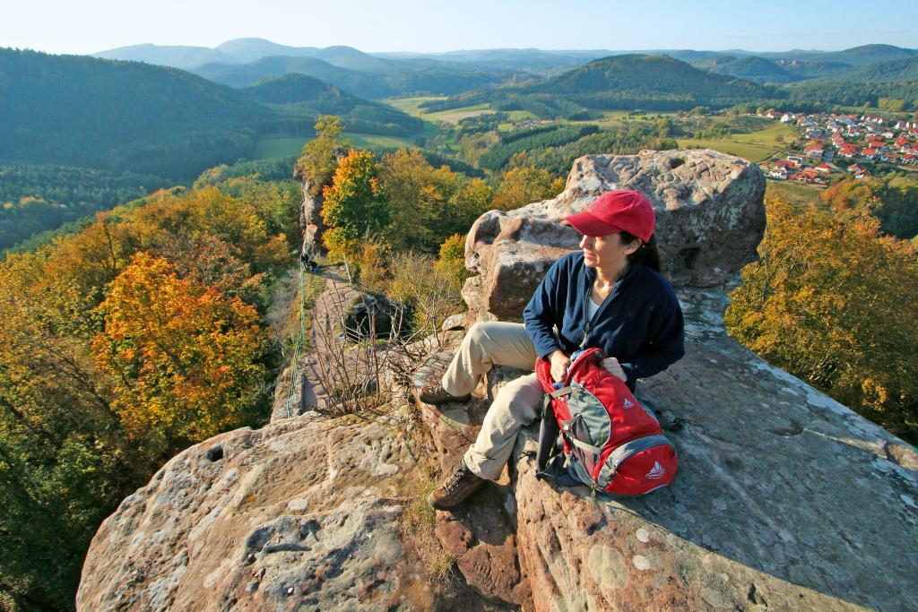 Eine Frau sitzt auf der Spitze eines hohen Felsens inmitten weiter, dicht bewaldeter Hügellandschaft, rechts im Hintergrund ein kleiner Ort mit rot gedeckten Häusern