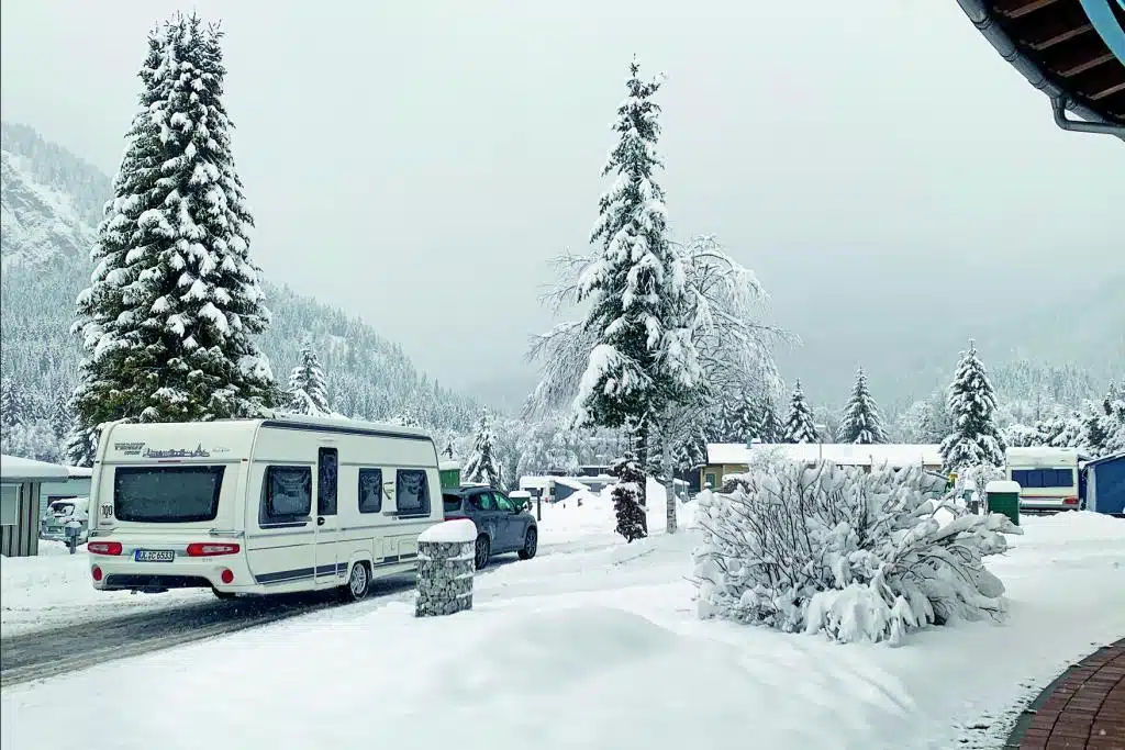 Ein Wohnwagen-Gespann fährt auf einem schnalen Weg über einen Campingplatz in den verschneiten Bergen.