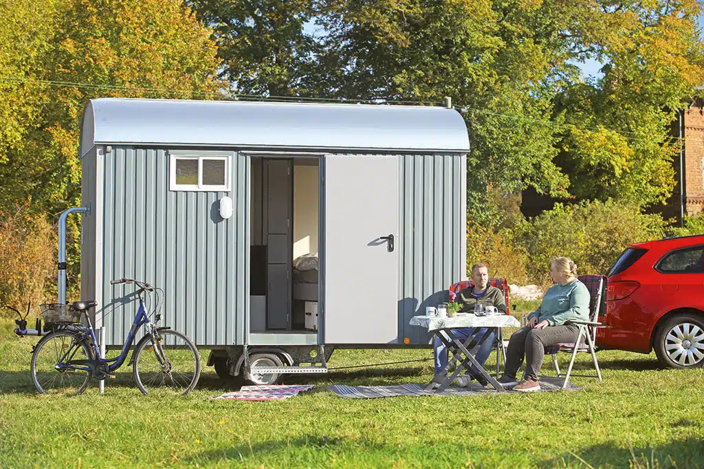 Kleiner Bauwagen auf einer Wiese mit Bäumen, vor dem Wagen sitzen eine Frau und ein Mann an einem Campingtisch.