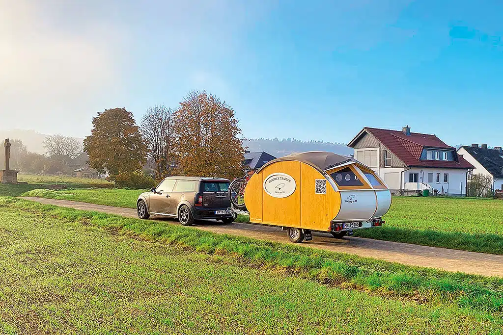 Miniwohnwagen in Teardrop-Form aus Holz hinter dunklem, kleinen PKW auf einer schmalen Straße zwischen Feldern, im Hintergrund ein Haus und einige Bäume