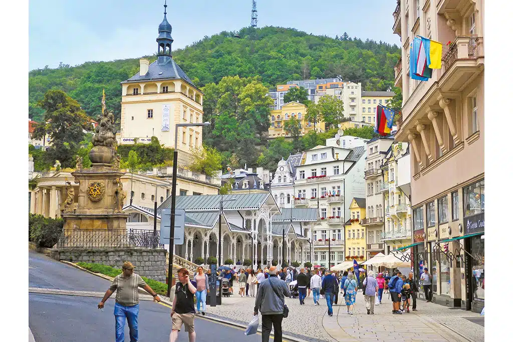 Eine Gasse mit vielen Passanten inmitten alter Stadtgebäude im Sommer, im Hintergrund erhebt sich ein bewaldeter Hügel.