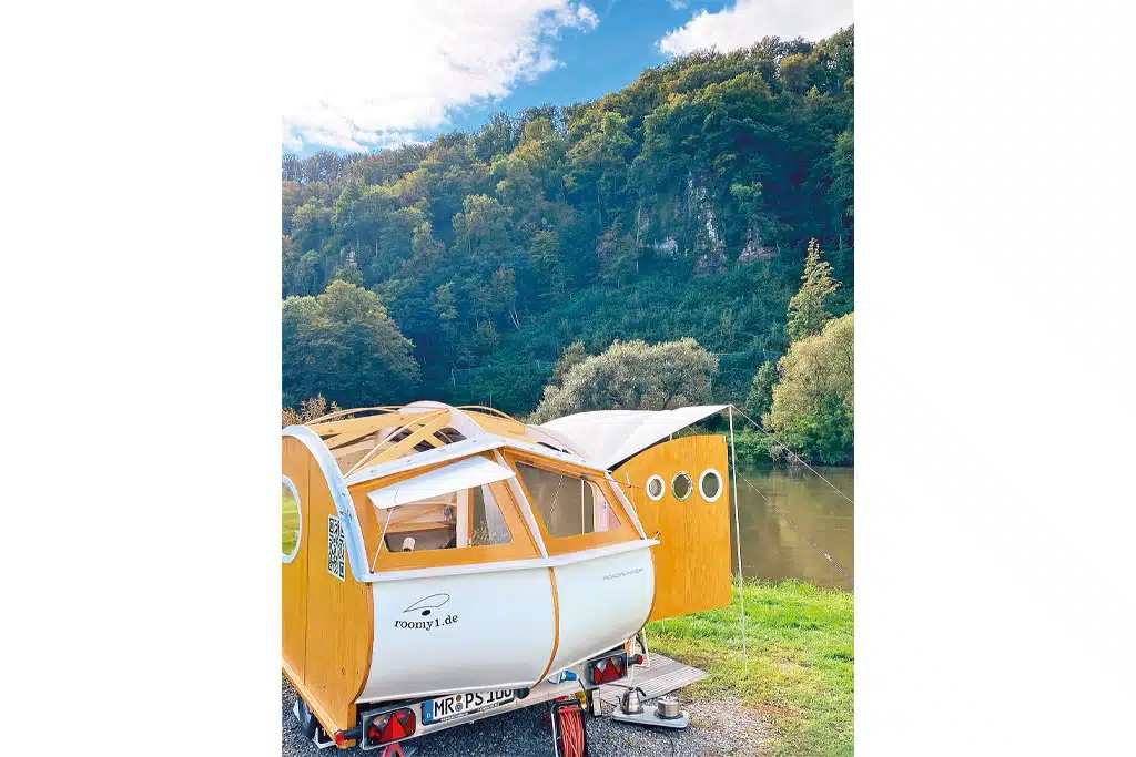 Kleiner Teardrop-Wohnwagen aus Holz mit offenem Dach und offener Flügeltür an einem Seeufer