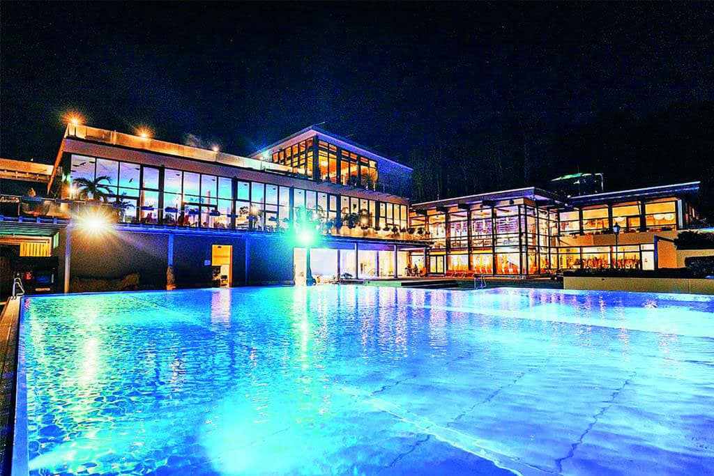 Ein beleuchteter Außenpool vor einem großen, beleuchteten Thermengebäude mit Fensterfronten bei Nacht