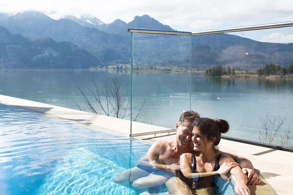 Eine Frau und ein Mann am Rand eines Außenpools, im Hintergrund ein See und eine Bergkette mit schneebedeckten Gipfeln