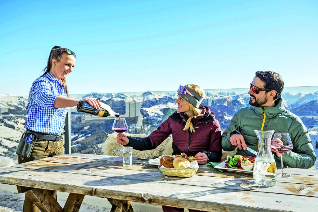 Eine Frau schenkt einer Frau und einem Mann an einem Holztisch einer Skihütte in den Bergen Wein ein.