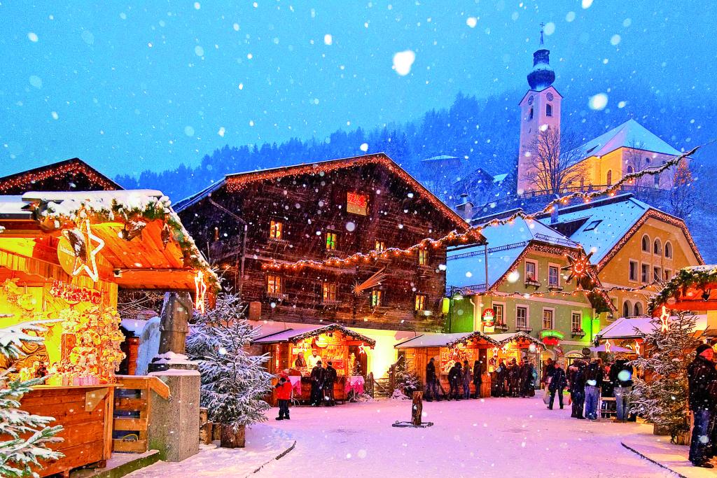 Einige beleuchtete Weihnachtsmarkt-Hütten vor einigen Gebäuden und einer Kirche in einem verschneiten Bergdorf