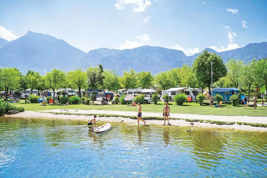 ADAC-Superplätze: Campingplatz an einem Seeufer mit Campingfahrzeugen und einigen Menschen am Wasser, im Hintergrund hohe Berge