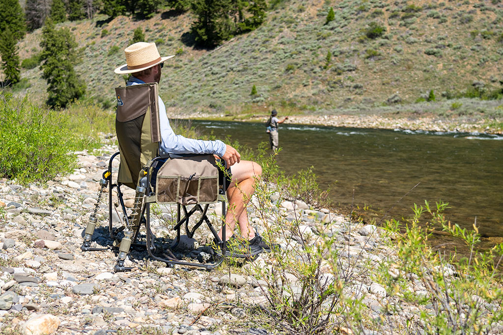 Mann sitzt auf einem Campingstuhl mit Federn von GCI Outdoor/SportTech, auf Kiesstrand vor einem Fluss.